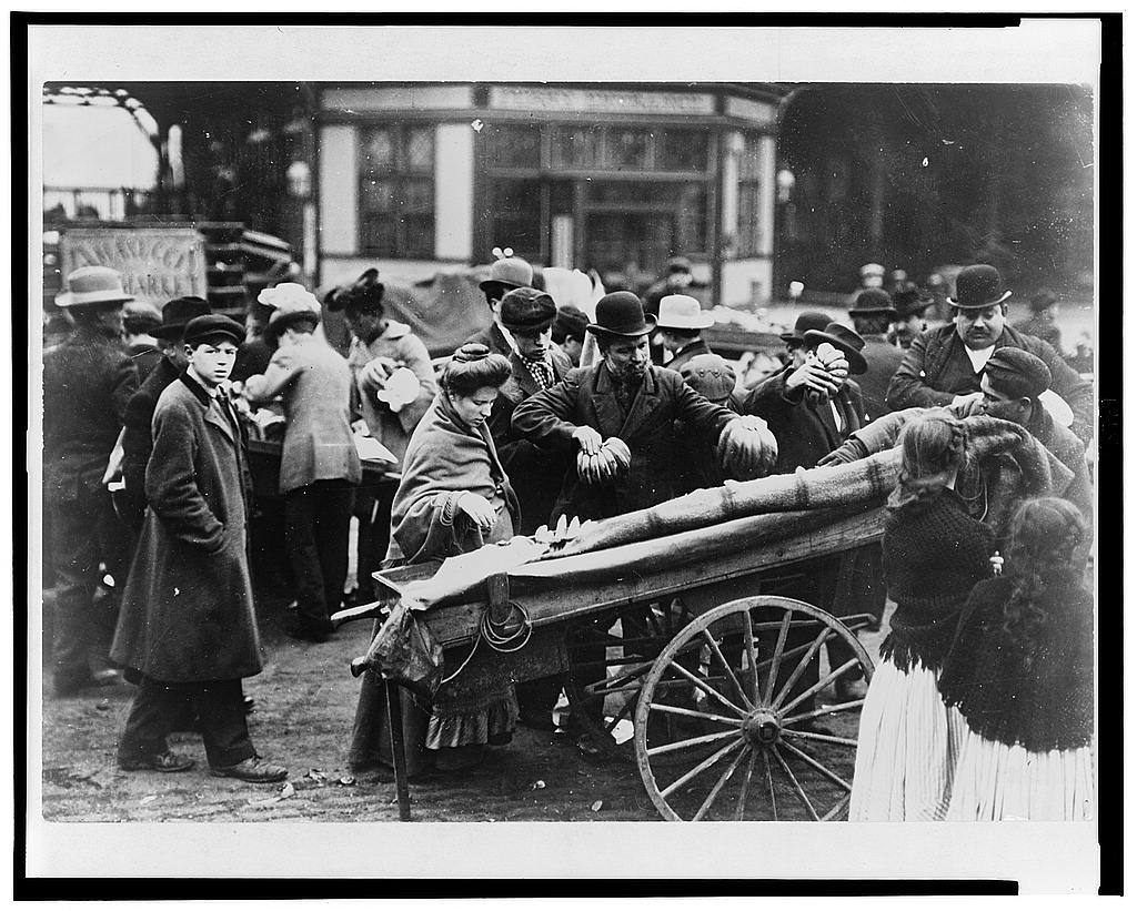 Italian immigrants composed of men, women and children are selecting bananas from a cart at an open-air market in Boston, MA’s Little Italy. An Italian immigrant man holds bananas in his hand while a woman peers into the cart. Young girls in shawls stand by. The street is crowded with people. The men wear coats and hats. It’s winter time.