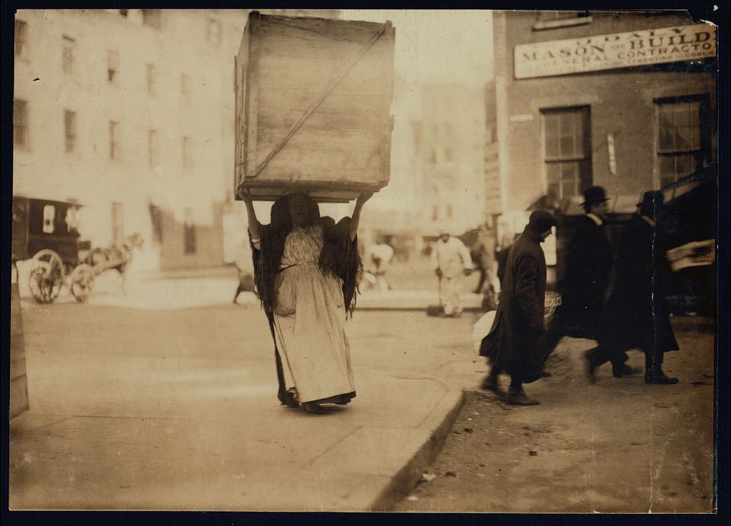Description: Italian woman carrying an enormous box above her head in New York City, NY. She wears a shawl that drapes over her, a dress and an apron. Men are nearby in coats and top hats. 